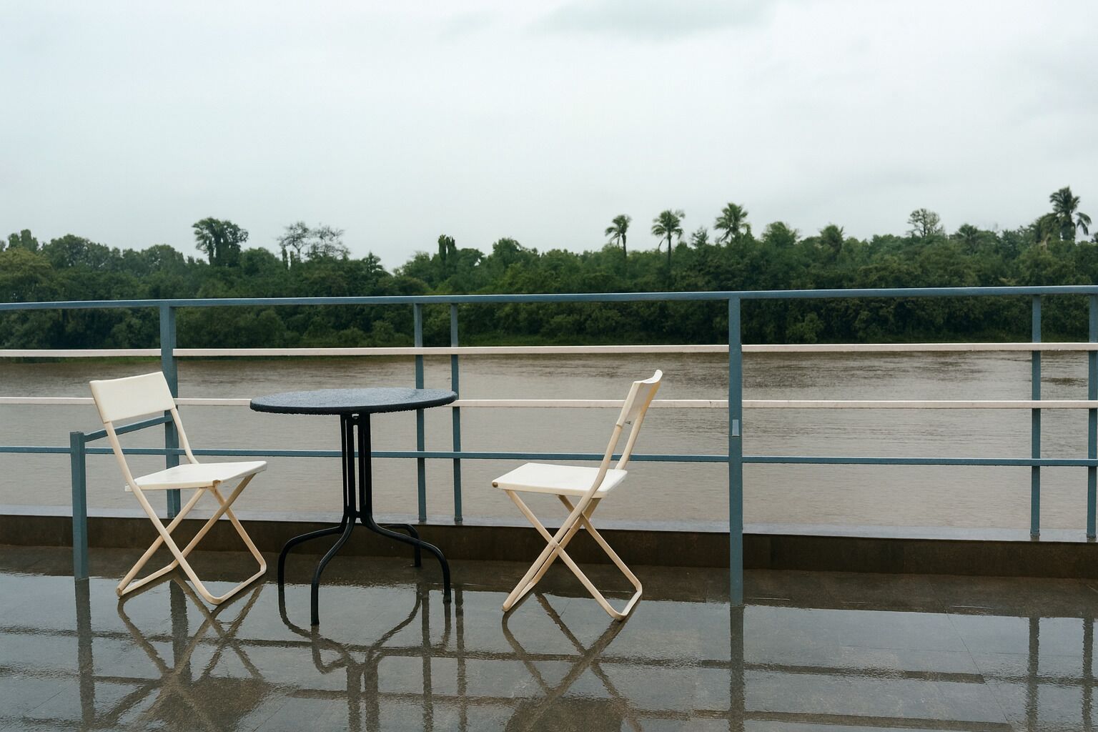 Twee witte klapstoelen en een kleine ronde tafel op een balkon met uitzicht op een rivier en groen.