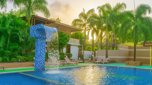 A swimming pool with a waterfall feature, surrounded by palm trees and lounge chairs. - River Splash Resort Neral Karjat (Karjat)