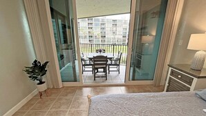 Ein Hotelzimmer mit einem Bett, einem kleinen Schreibtisch, einer Zimmerpflanze und einer Glastür, die auf einen Balkon mit Tisch und Stühlen führt.