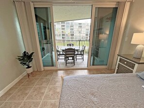 A hotel room with a bed, a small desk, a potted plant, and a sliding glass door leading to a balcony with a table and chairs. - Captains Quarters Oceanfront – Saltwater Sunrises (St. Augustine)