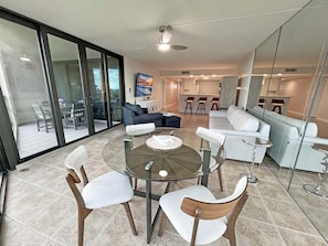 A modern living room with a glass dining table, white sofas, and a kitchen area in the background.