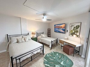 Ein Schlafzimmer mit zwei Betten, einem Schreibtisch, einem Stuhl, einem Deckenventilator und Kunstwerken an den Wänden.