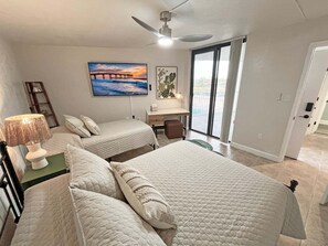 A hotel room with two beds, a desk, a chair, a lamp, and a view of the outdoors. - Captains Quarters Oceanfront – Saltwater Sunrises (St. Augustine)