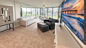 Ein geräumiges Wohnzimmer mit großem Fenster, einem weißen Sofa, einem schwarzen Sofa und einem Gemälde mit Sonnenuntergang über dem Meer.