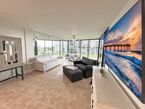 A spacious living room with a large window, a white sofa, a black sofa, and a painting of a sunset over the ocean. - Captains Quarters Oceanfront – Saltwater Sunrises (St. Augustine)