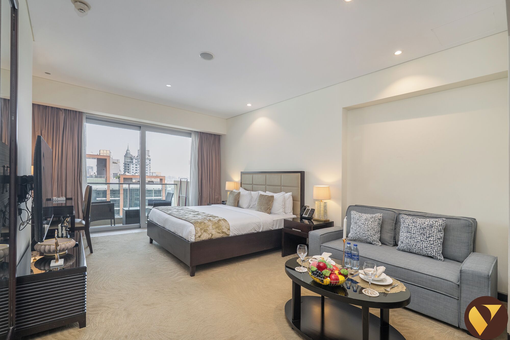 Une chambre d’hôtel avec un grand lit, un canapé, une table à manger avec des fruits et des bouteilles d’eau, et une vue sur la ville par la fenêtre.