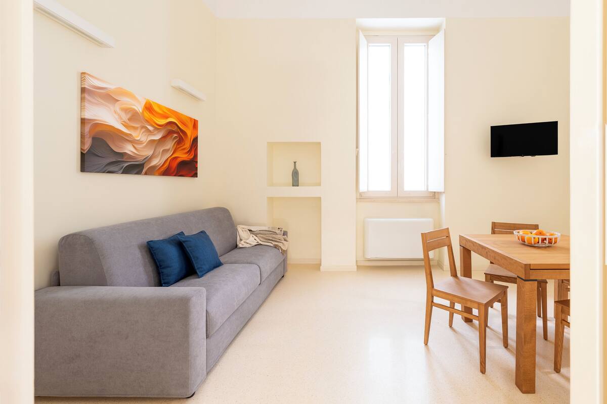 A modern living room with a grey sofa, a wooden dining table, and a TV mounted on the wall.