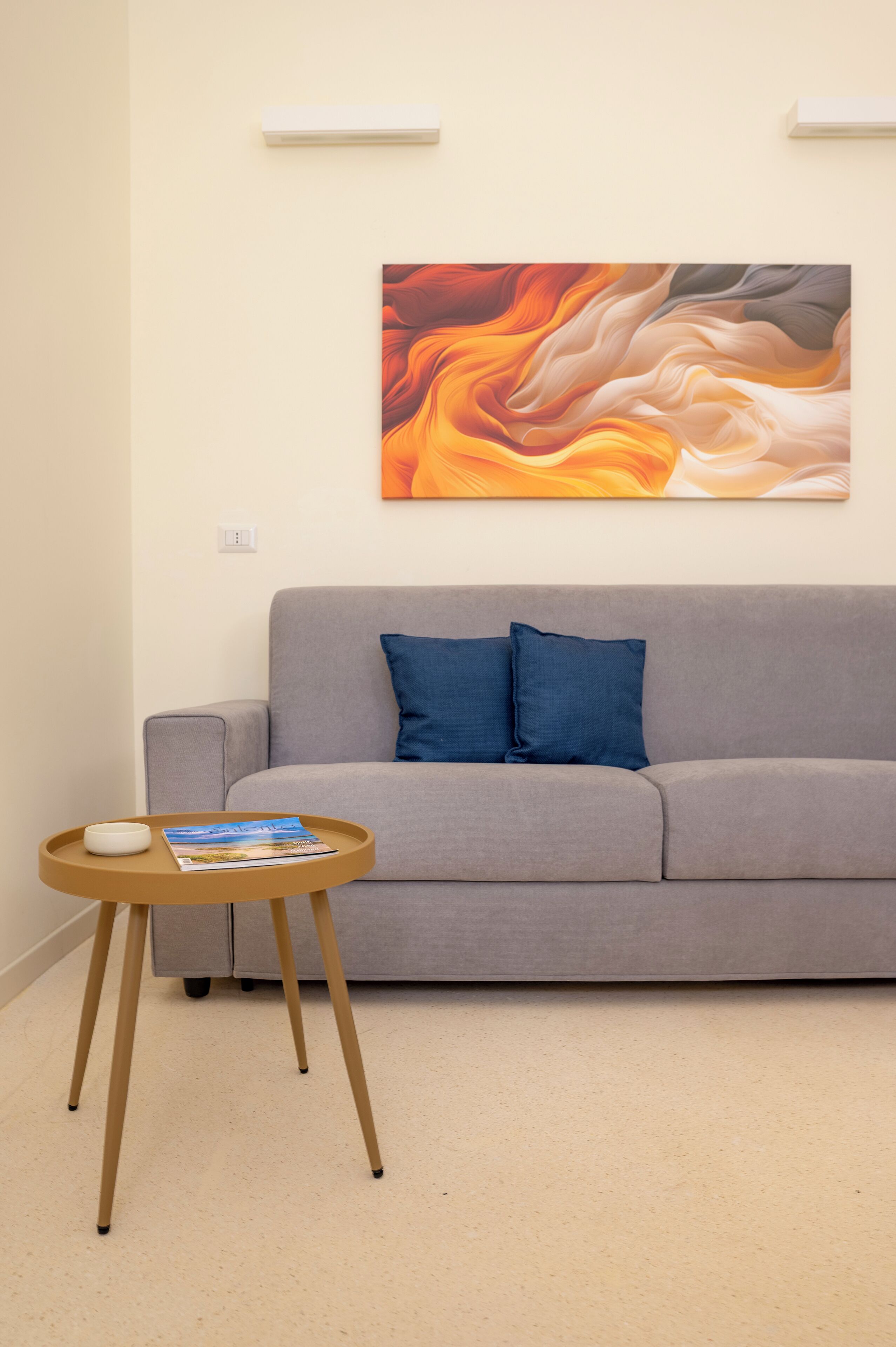 Un soggiorno moderno con un divano grigio, due cuscini blu, un tavolino di legno con una rivista e un grande quadro astratto appeso al muro.