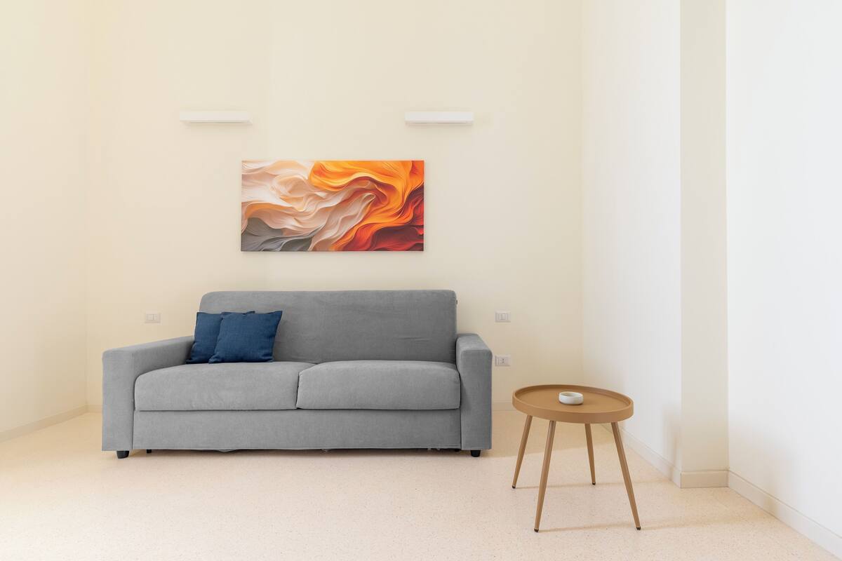 A modern living room with a grey sofa, a small round wooden side table, and a colorful abstract painting on the wall.
