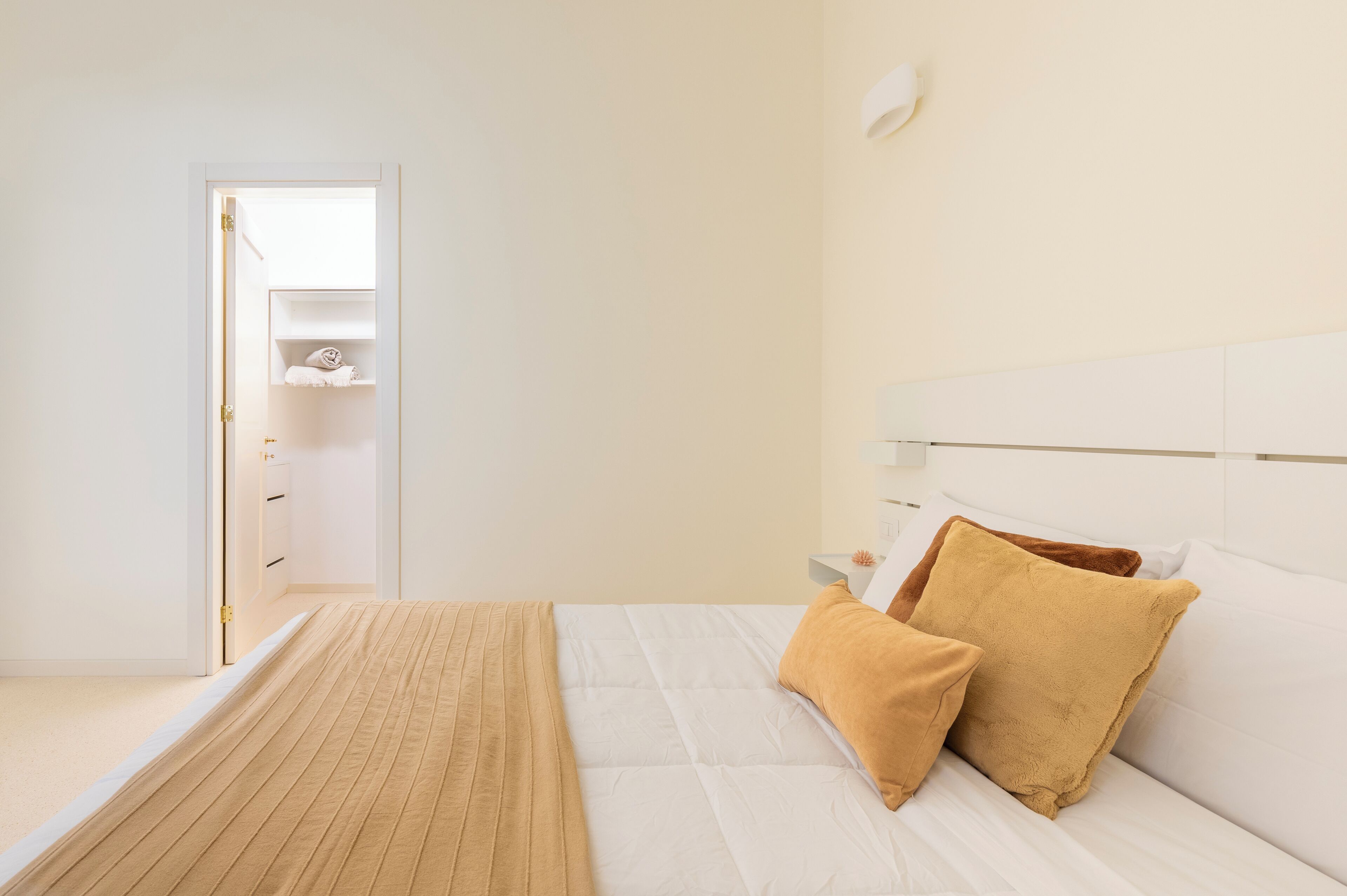 A bedroom with a bed, a bedside table, a nightstand, a wall-mounted lamp, and a door leading to a bathroom.