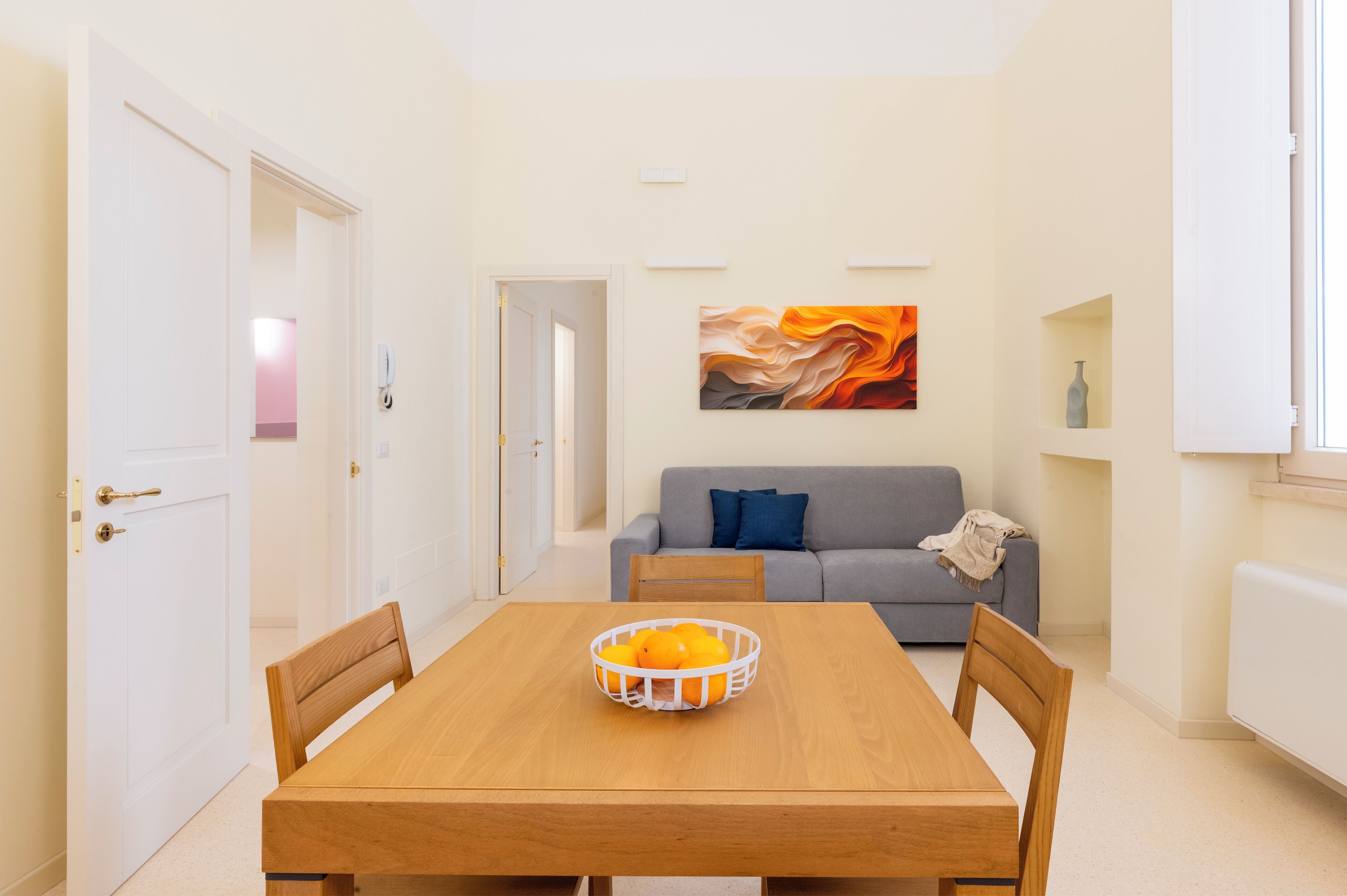 Un coin repas avec une table en bois, des chaises et un saladier rempli d’oranges. On aperçoit, à l’arrière-plan, un canapé gris avec un coussin bleu.
