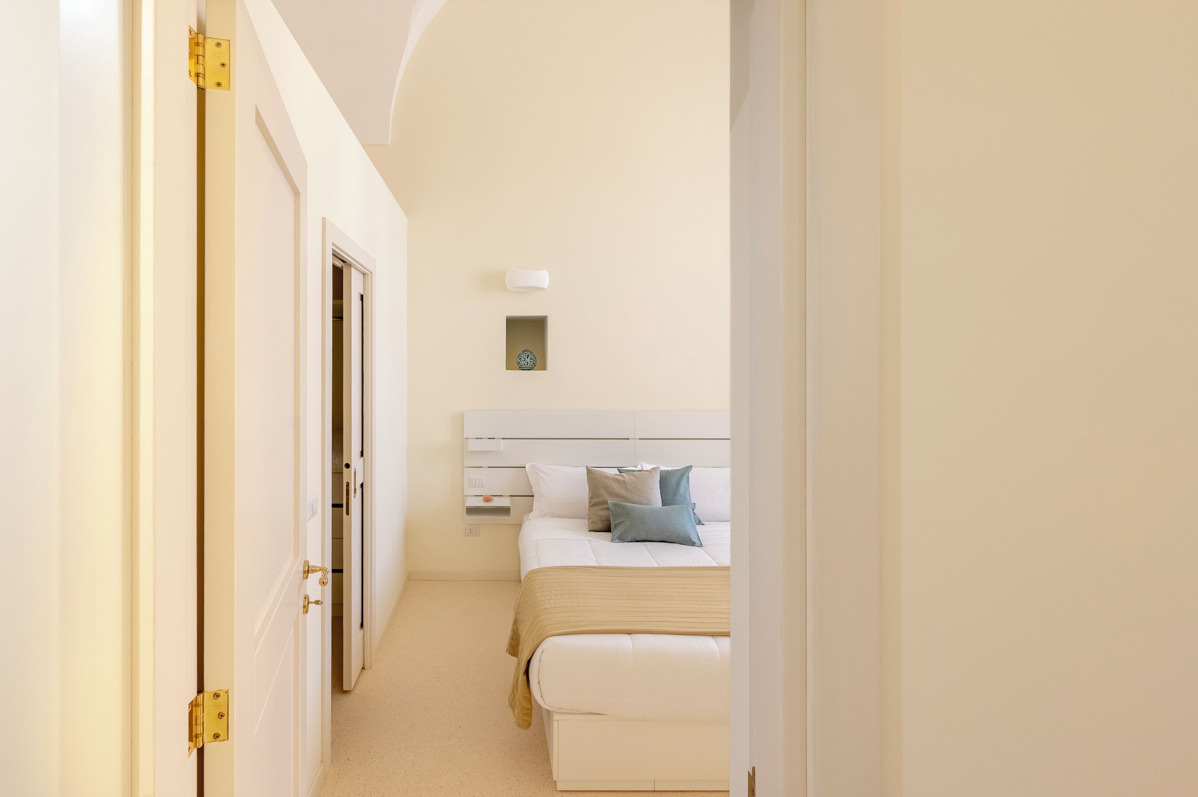 Een smalle gang die leidt naar een slaapkamer met een bed, nachtkastje en een klein wandlampje.