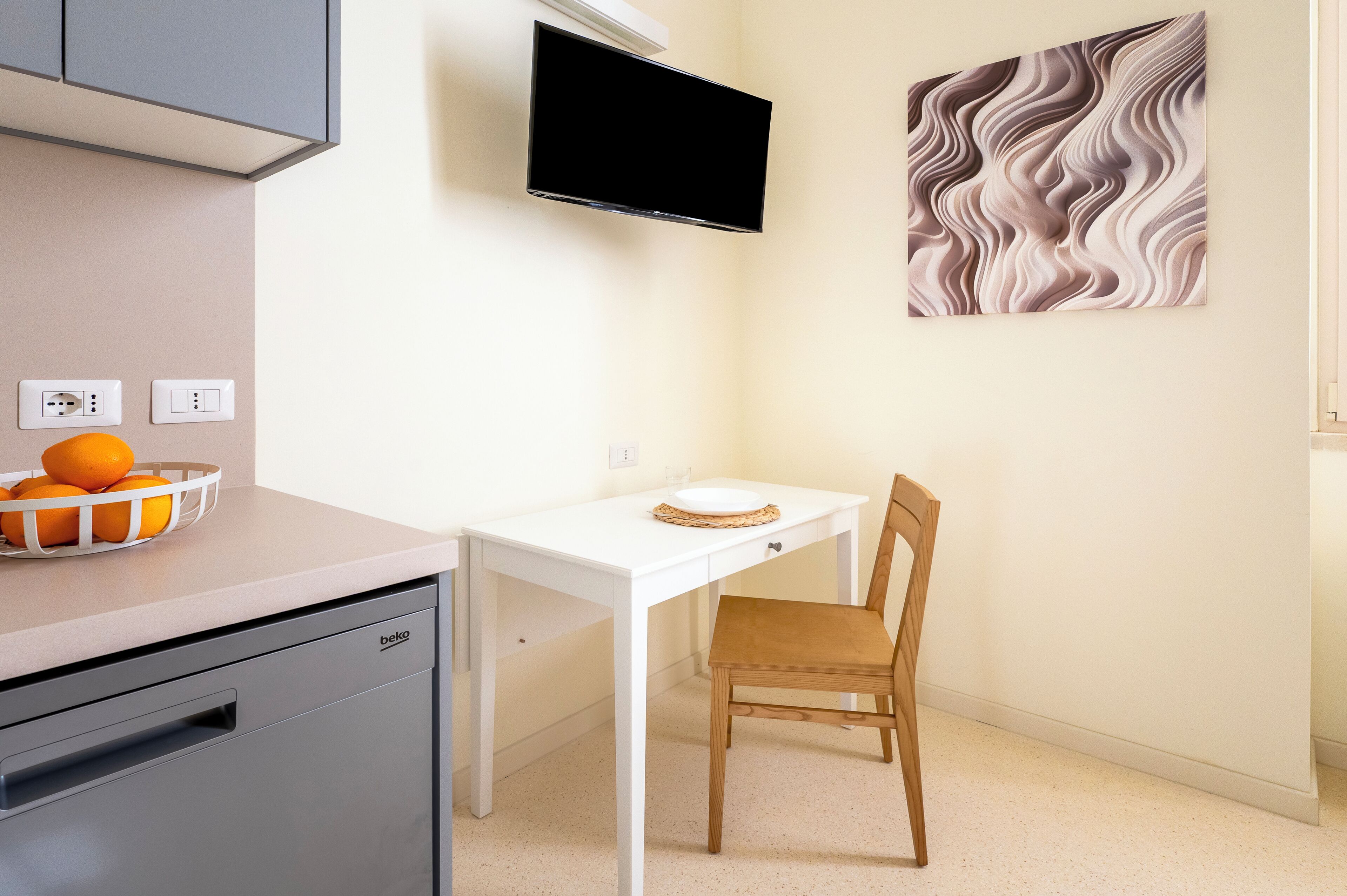A kitchen with a white countertop, a sink, a dish rack with oranges, a white table with a chair, a flat-screen TV, and a wall-mounted artwork.