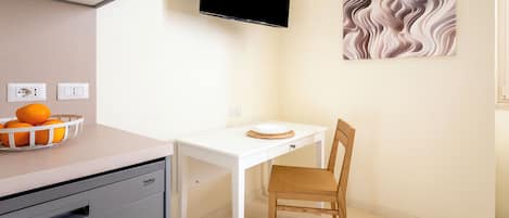 A kitchen with a white countertop, a sink, a dish rack with oranges, a white table with a chair, a flat-screen TV, and a wall-mounted artwork.
