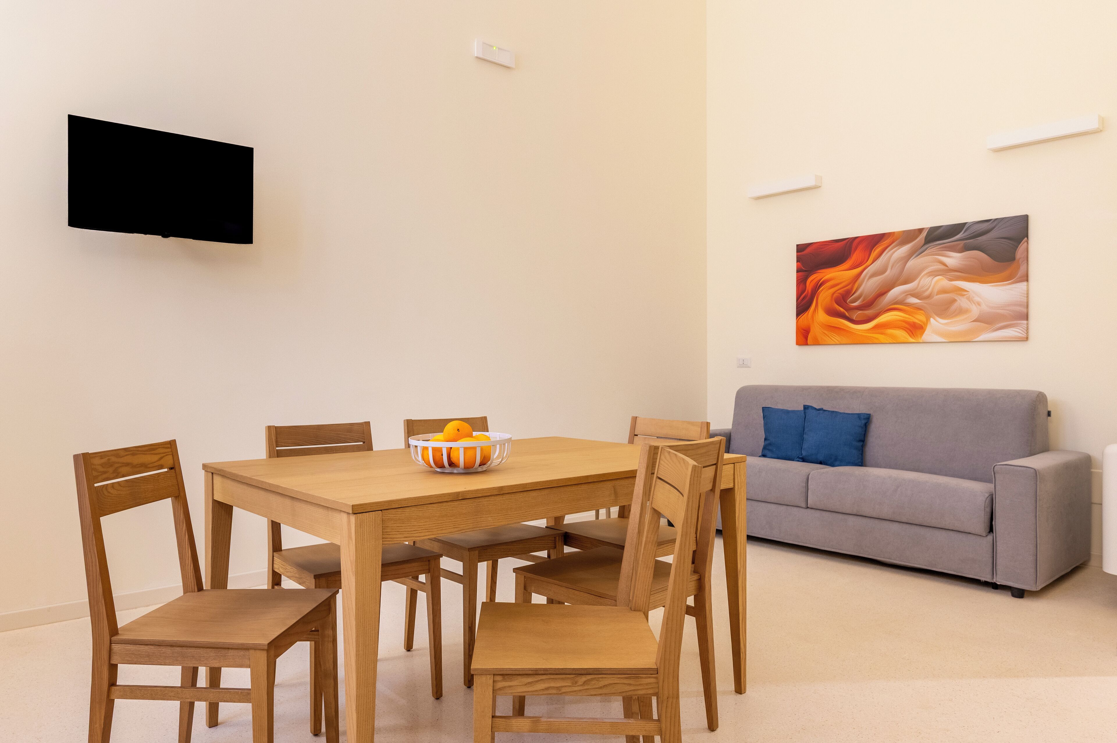 A dining area with a wooden table and chairs, a bowl of fruit, a grey sofa, and a flat-screen TV mounted on the wall.