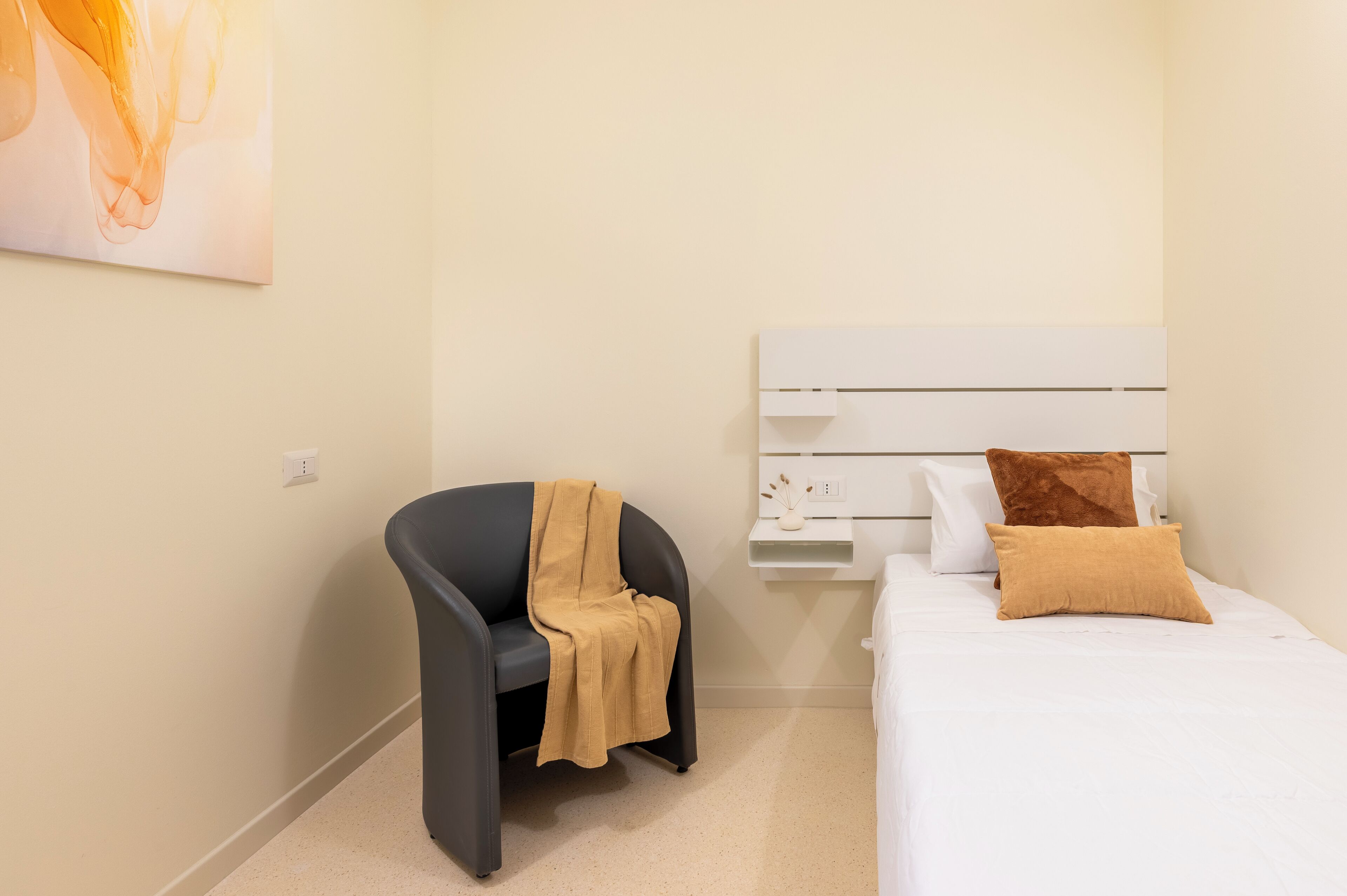 A bedroom with a bed, a chair, a nightstand, and a wall-mounted shelf.