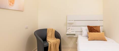 A bedroom with a bed, a chair, a nightstand, and a wall-mounted shelf.