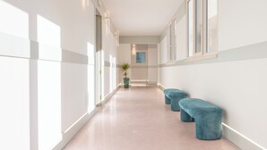 A hallway with white walls, large windows, and a plant. There are two blue benches along the wall. - Lùmia Lecce - Urban Residence (Lecce)