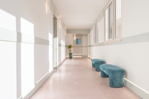 Un pasillo con paredes blancas, ventanas amplias y una planta. Hay dos bancos azules a lo largo de la pared.