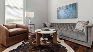 Una sala de estar con un sofá de cuero marrón, un sofá capitonado gris, una mesa de centro redonda, una lámpara blanca y un cuadro abstracto grande en la pared.