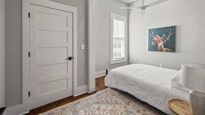 Un dormitorio con una puerta blanca, una cama con ropa de cama blanca, un ventilador de techo, una ventana con persianas y un cuadro de un ciervo.