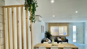 A modern living room with a wooden dining table, white chairs, a sofa, and a large window.