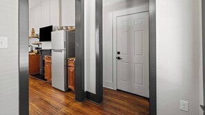 Een moderne keuken met witte kastjes, een koelkast en een magnetron. Er ligt een houten vloer en er is een deur naar een andere kamer.