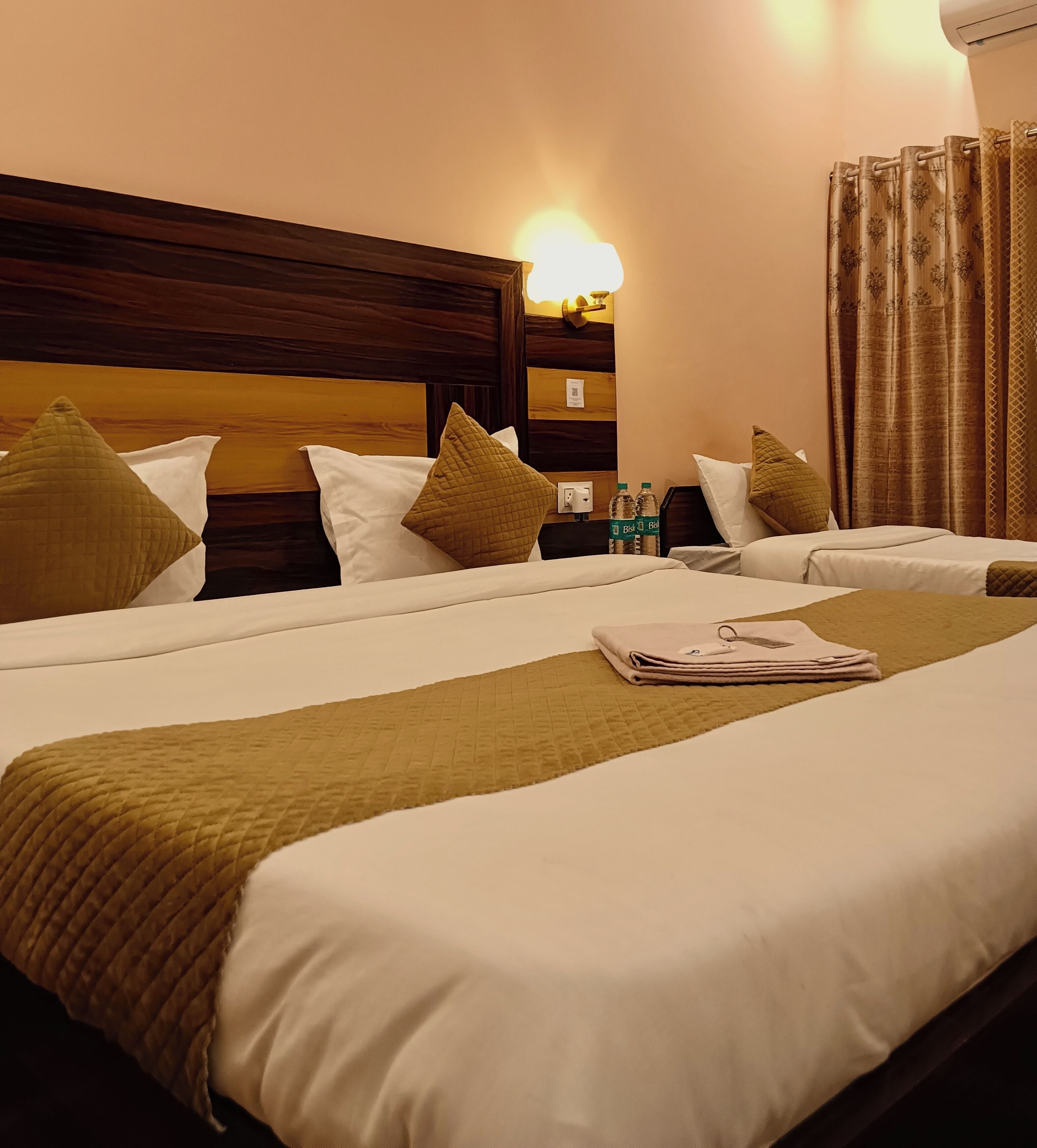 Habitación de hotel con dos camas, cada una con cabecero de madera, ropa de cama blanca y almohadones verdes. Se ve un mesita de noche con botellas de agua y una lámpara.