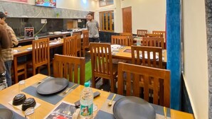 A restaurant with wooden tables and chairs, a ceiling fan, and a kitchen area in the background. - Geetanjali Palace  (Jakholi)