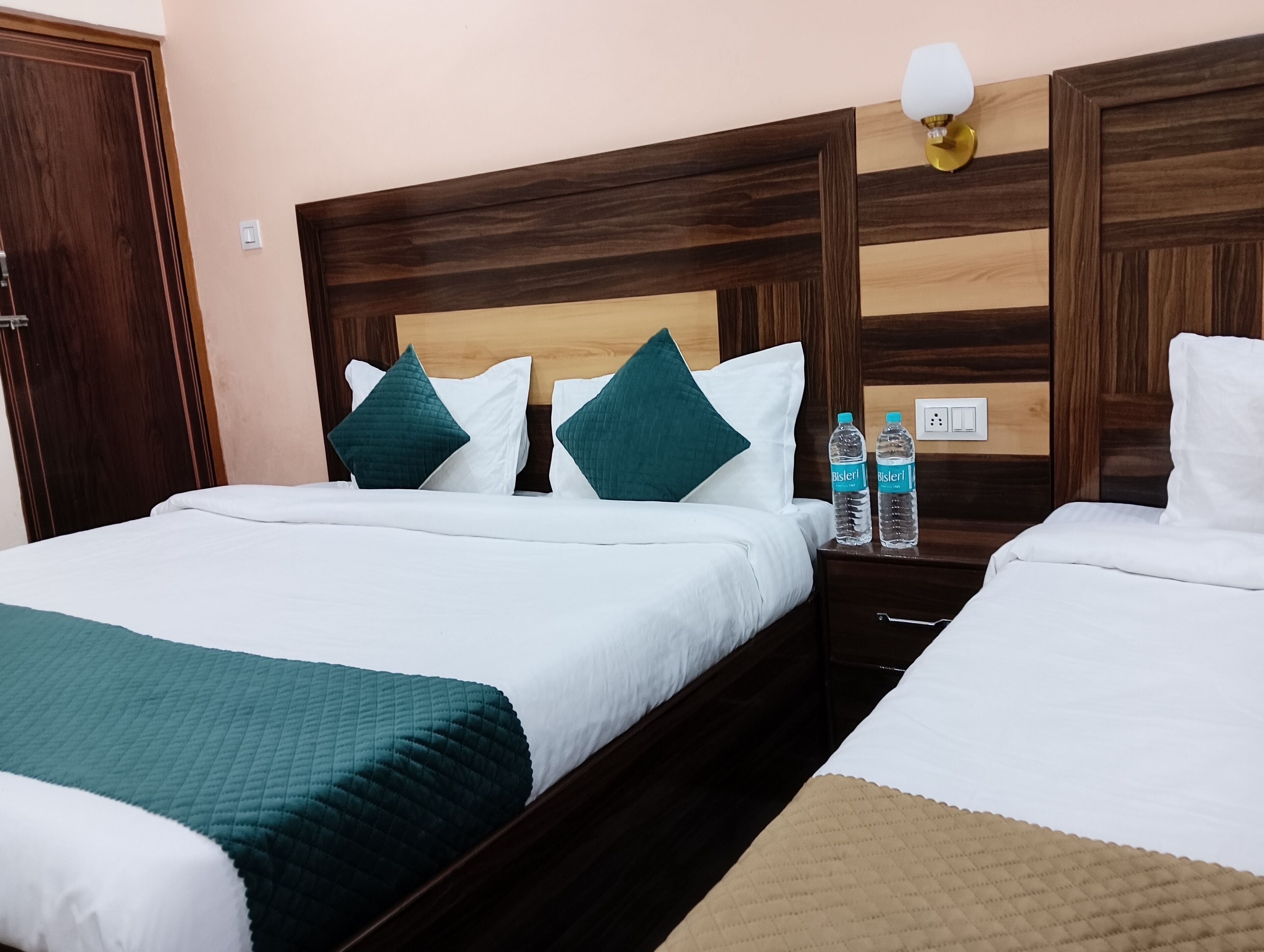 A hotel room with two beds, each with white bedding and teal pillows. There are two water bottles on the nightstand between the beds.