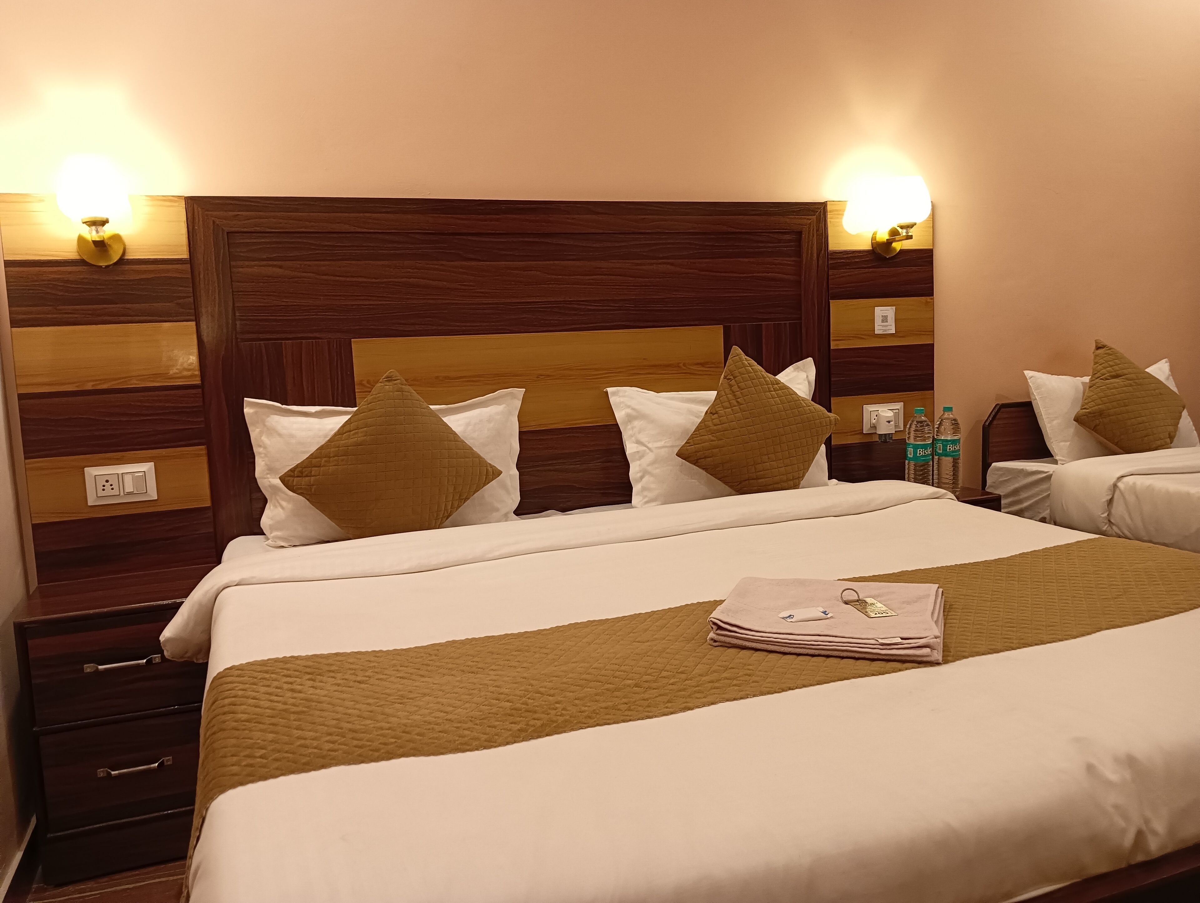 Habitación de hotel con una cama grande, dos mesitas de noche y un cabecero de madera. Hay dos lámparas de pared y una botella de agua en la mesita.