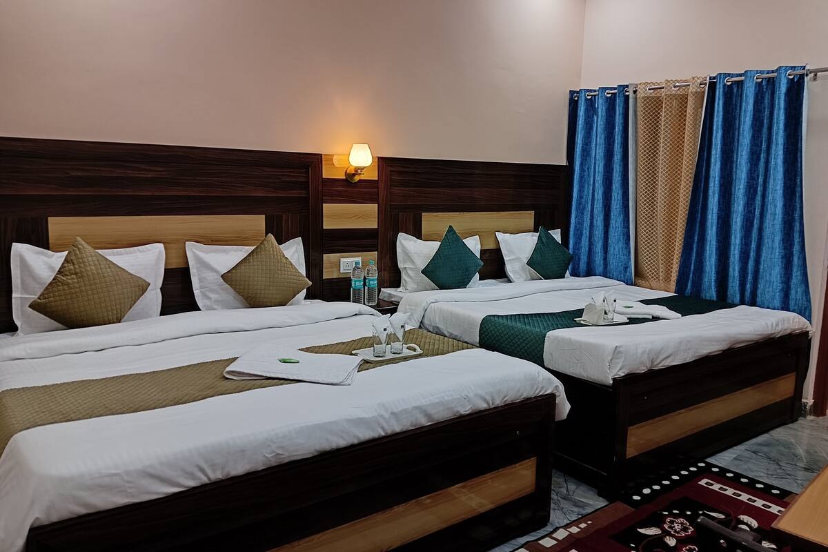 Ein Hotelzimmer mit zwei Betten, Holz-Kopfteilen, weißer Bettwäsche, grün-beigen Kissen, einem Nachttisch mit Wasserflaschen, einer Lampe und einem gemusterten Teppich.