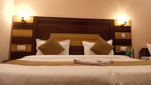 A neatly made bed with a wooden headboard, two wall-mounted light fixtures, and a small table with water bottles beside it. - Geetanjali Palace  (Jakholi)