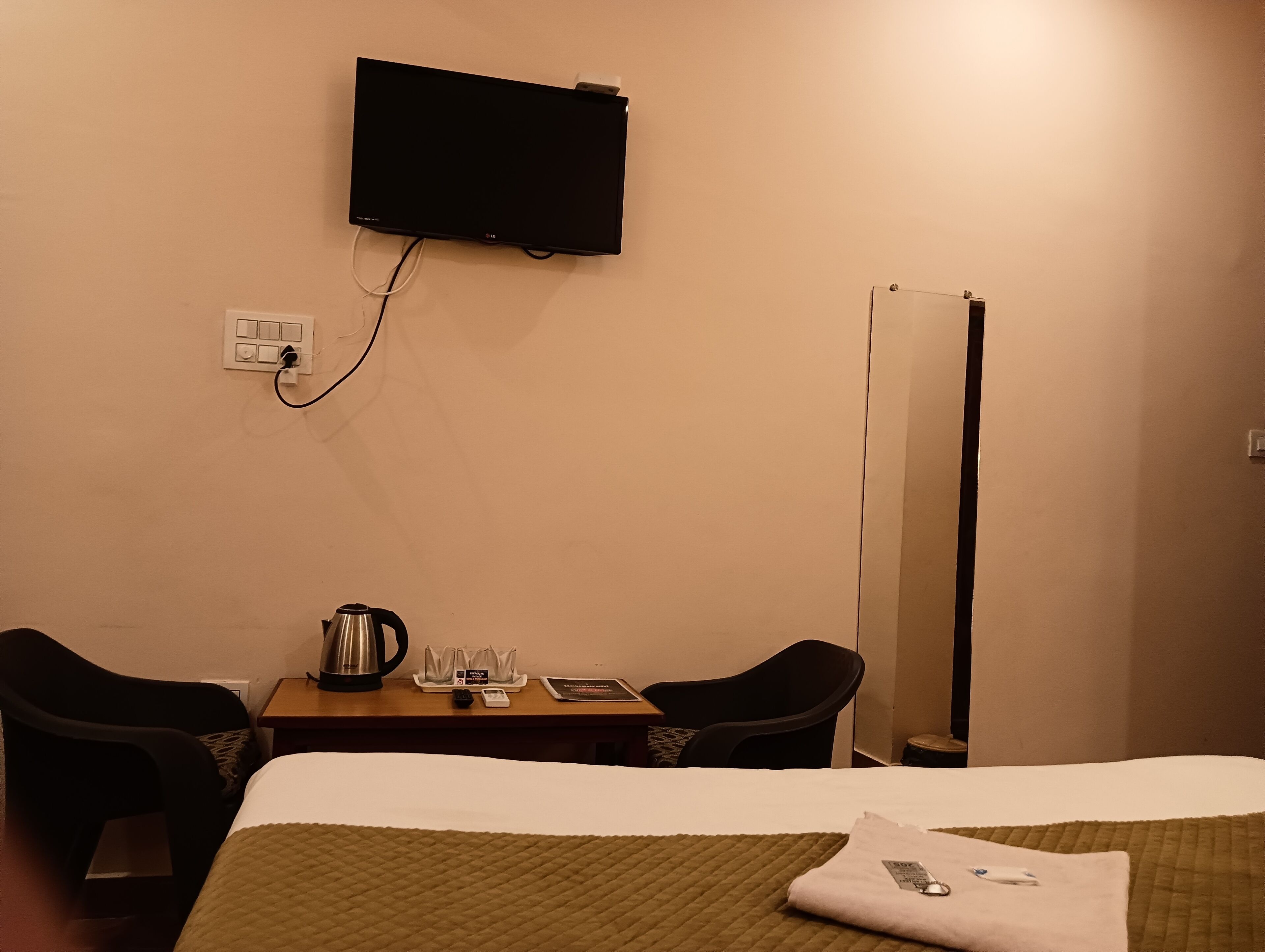 A hotel room with a bed, a desk with a kettle, a flat-screen TV mounted on the wall, and a mirror.