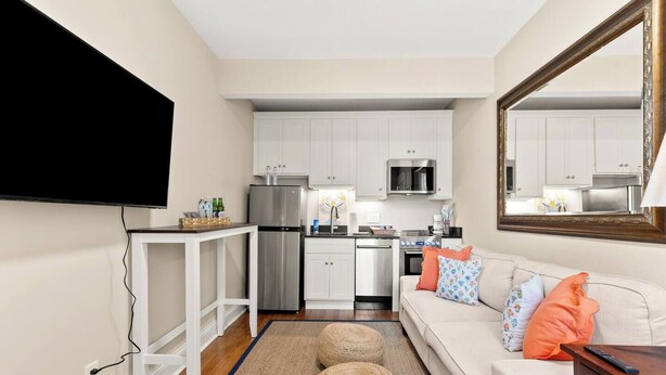 A compact living space with a kitchenette, a flat-screen TV mounted on the wall, a sofa with patterned cushions, and a large mirror.
