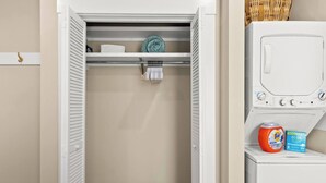 A laundry room with a washing machine and a dryer, a shelf with folded towels, and a basket of laundry supplies.
