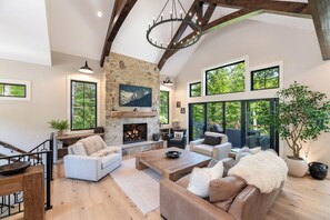 A spacious living room with a stone fireplace, wooden beams, and large windows offering a view of the outdoors.