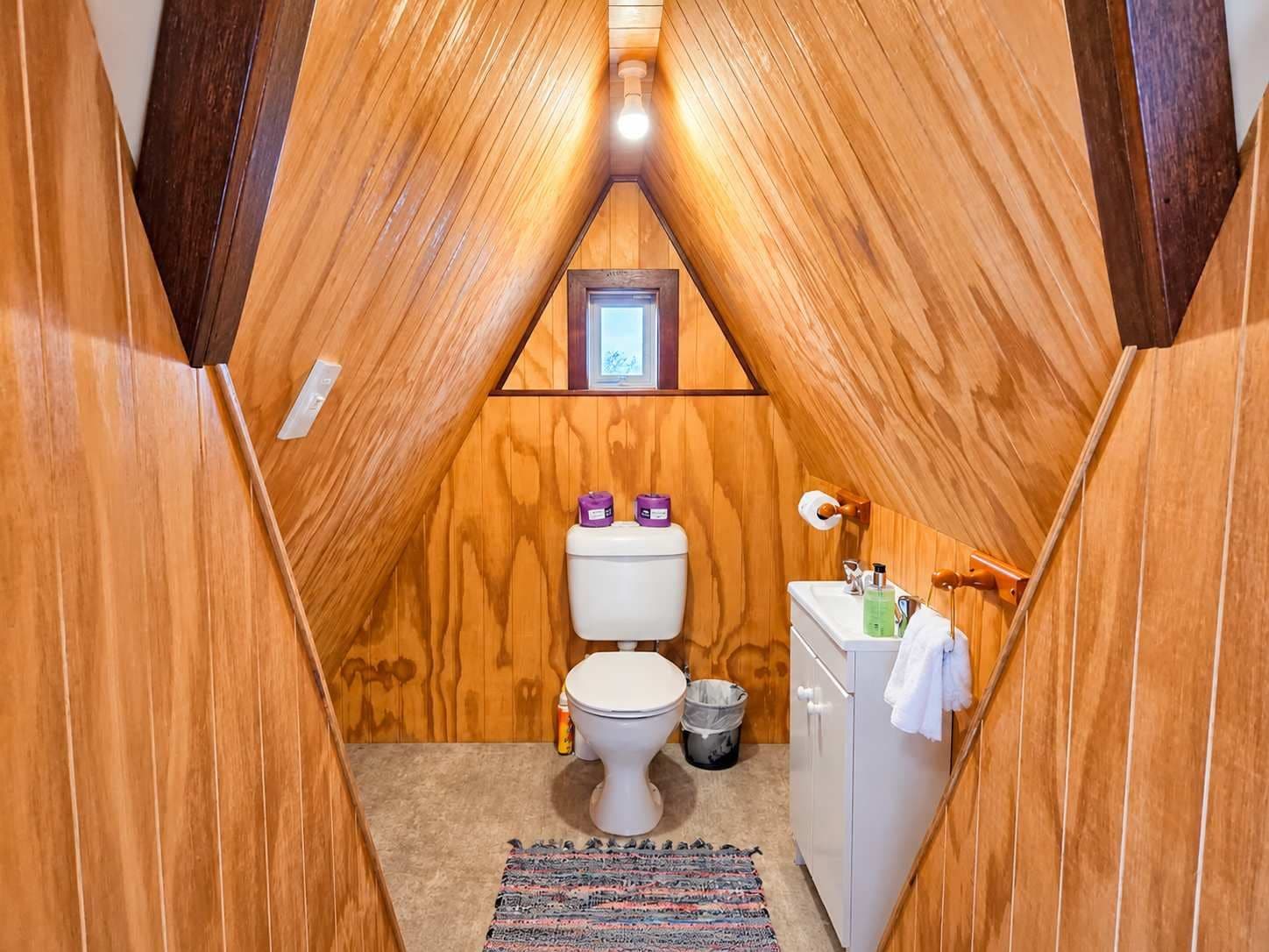 A small, wooden-paneled bathroom with a white toilet, a sink, and a trash bin.