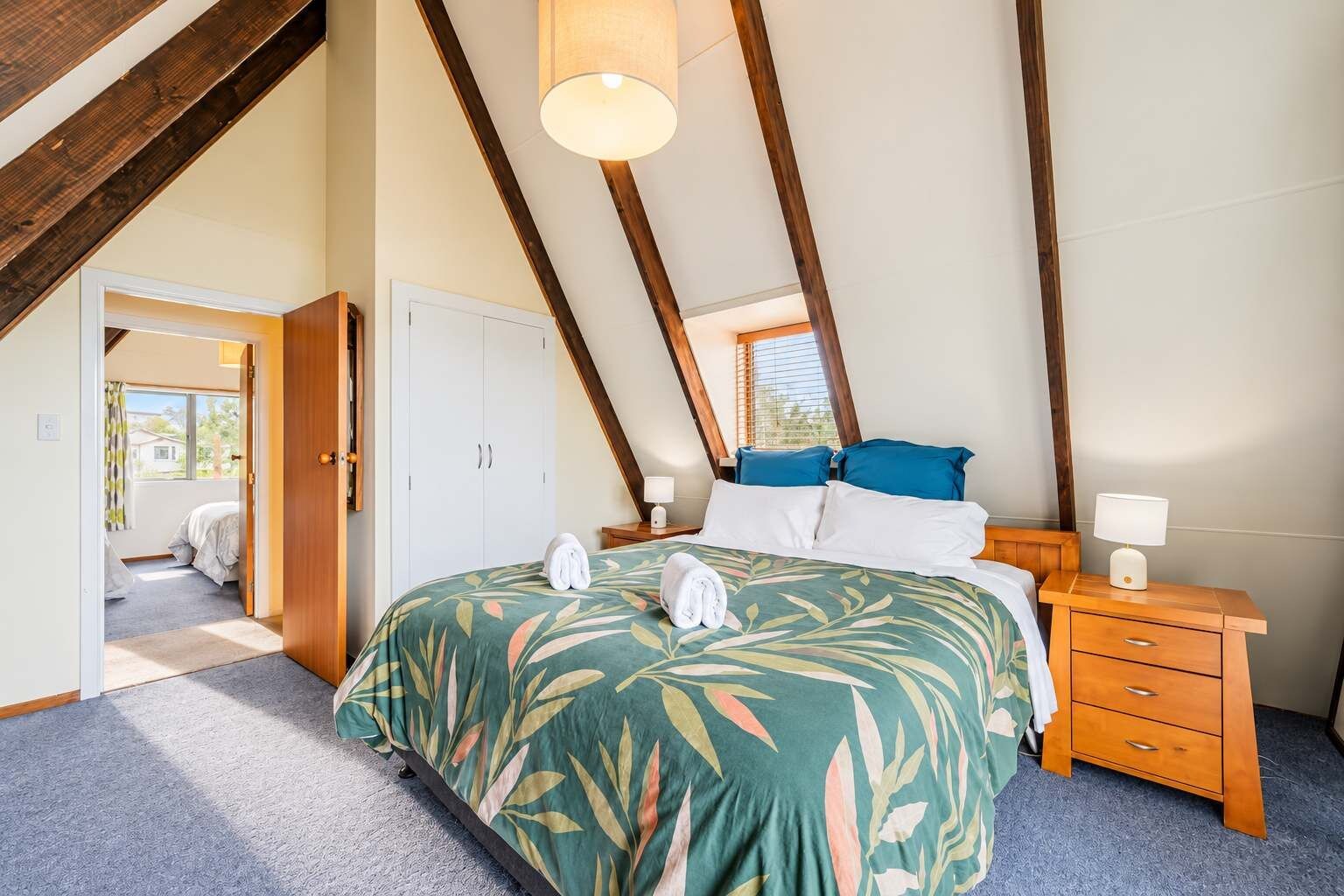 A bedroom with a sloped ceiling, a bed with a floral bedspread, a nightstand with a lamp, and a door leading to another room.
