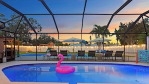 A swimming pool with a pink flamingo float, surrounded by lounge chairs and umbrellas, under a covered area with string lights.