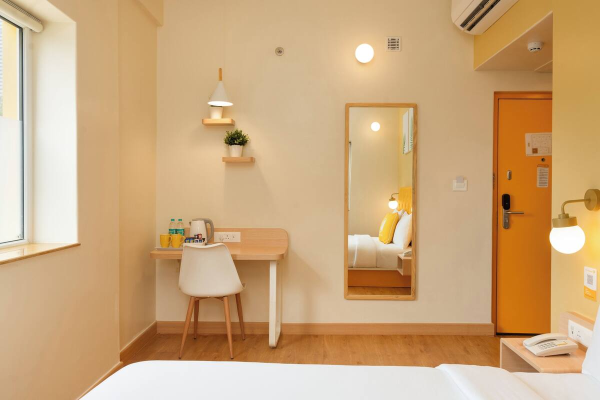 Habitación de hotel con cama, escritorio con silla, espejo, mesita de noche con teléfono y puerta con lámpara.