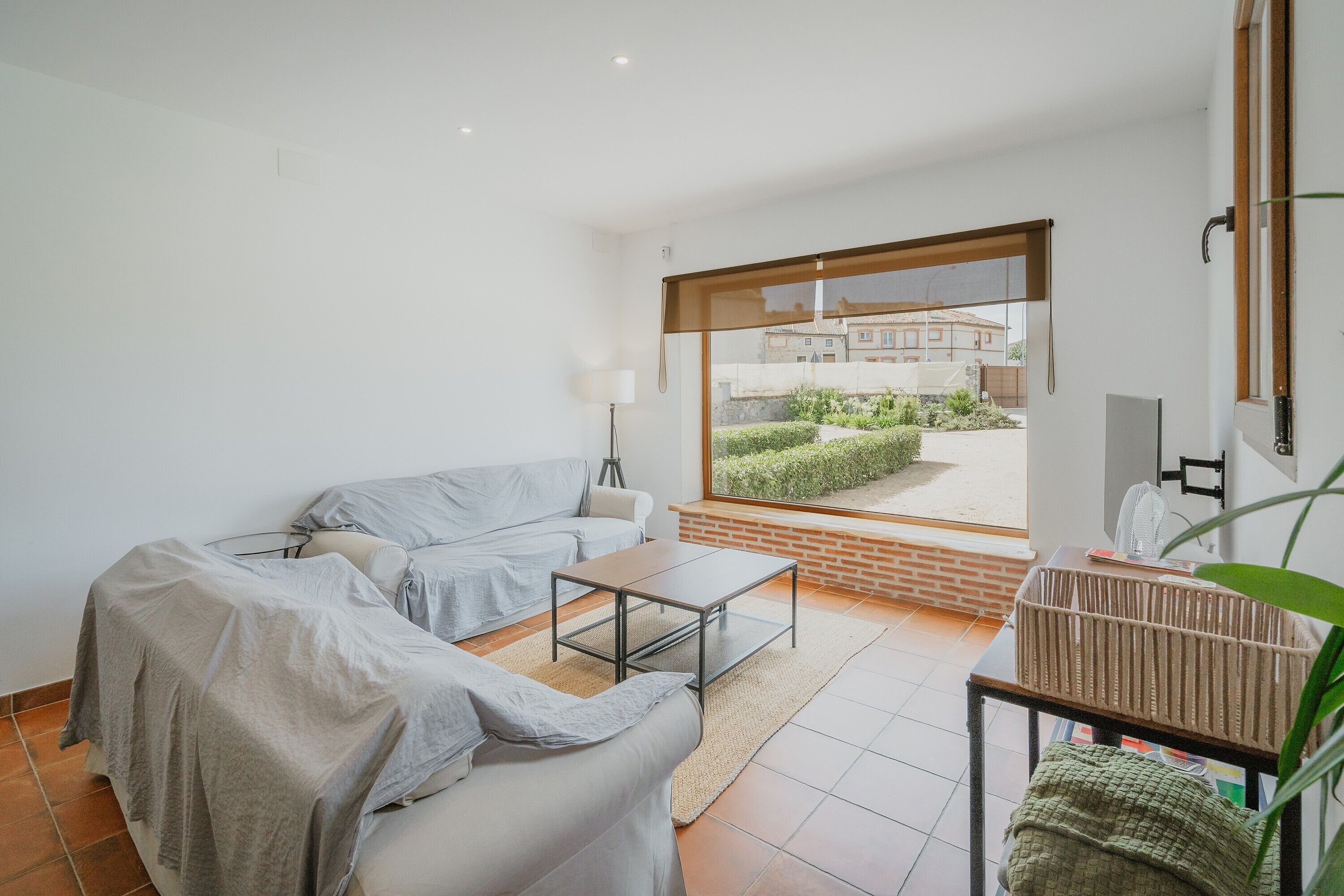 Un salon moderne comprenant un canapé, une table basse, une lampe et une grande fenêtre donnant sur un jardin et des immeubles.