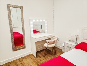 A bedroom with a vanity, a mirror with lights, a chair, and a bed with red bedding. - Cleave House - Stylish n Chic 3 Bed House Mins From Central London (Orpington)