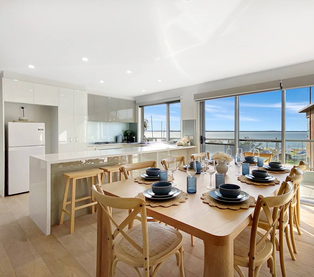 Une cuisine et une salle à manger modernes, dotées d’un grand îlot central, de chaises en bois et offrant une vue sur la mer.