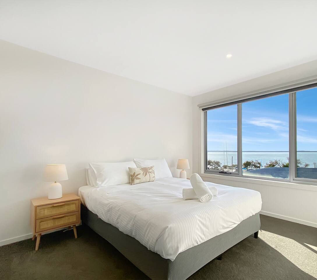 A bedroom with a large window, a bed, a nightstand, and a lamp.