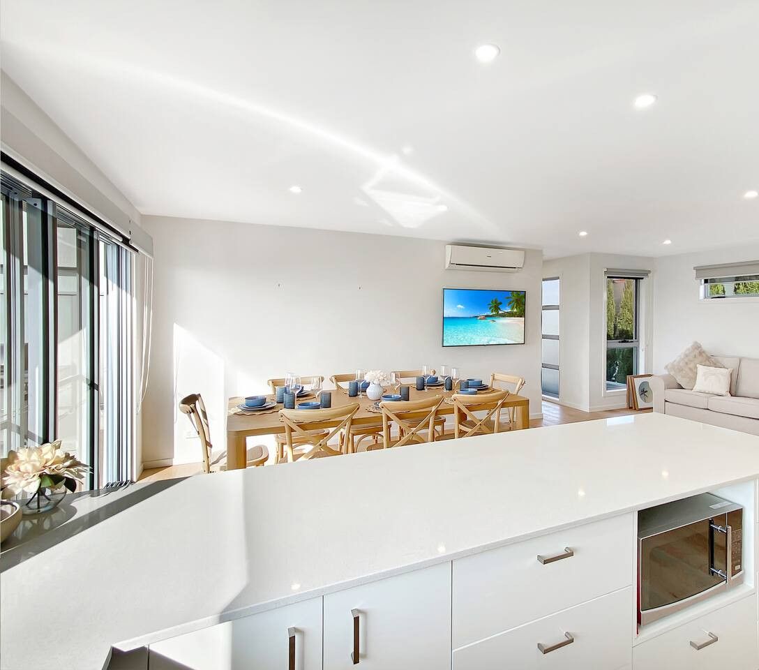 A modern kitchen with a central island, a dining area with a view of the sea, and a living area with a sofa and cushions.