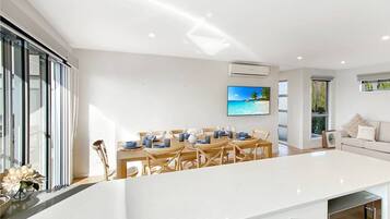 A modern kitchen with a central island, a dining area with a view of the sea, and a living area with a sofa and cushions.
