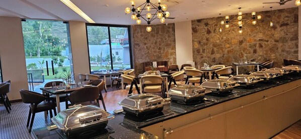 A buffet setup with multiple serving stations, a stone accent wall, and a view of the outdoors through large windows. - Ullasa by Momentum (Udaipur)