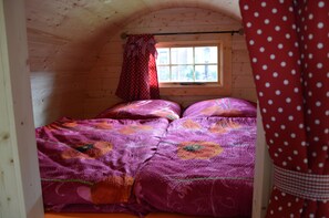 Une pièce chaleureuse aux murs en bois, avec une fenêtre ornée de rideaux et un lit recouvert d’une couvre-lit à motifs floraux.