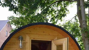 Een houten, cilindervormig gebouw met een open deur, een bankje ervoor en een bed binnen.